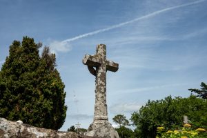 A weathered stone cross stands in a Breton cemetery against a vibrant blue sky, surrounded by lush greenery.